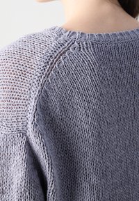 Suéter de punto gris claro con tejido suelto, con cuello acanalado y panel trasero texturizado. Muestra costuras detalladas y un ajuste relajado.