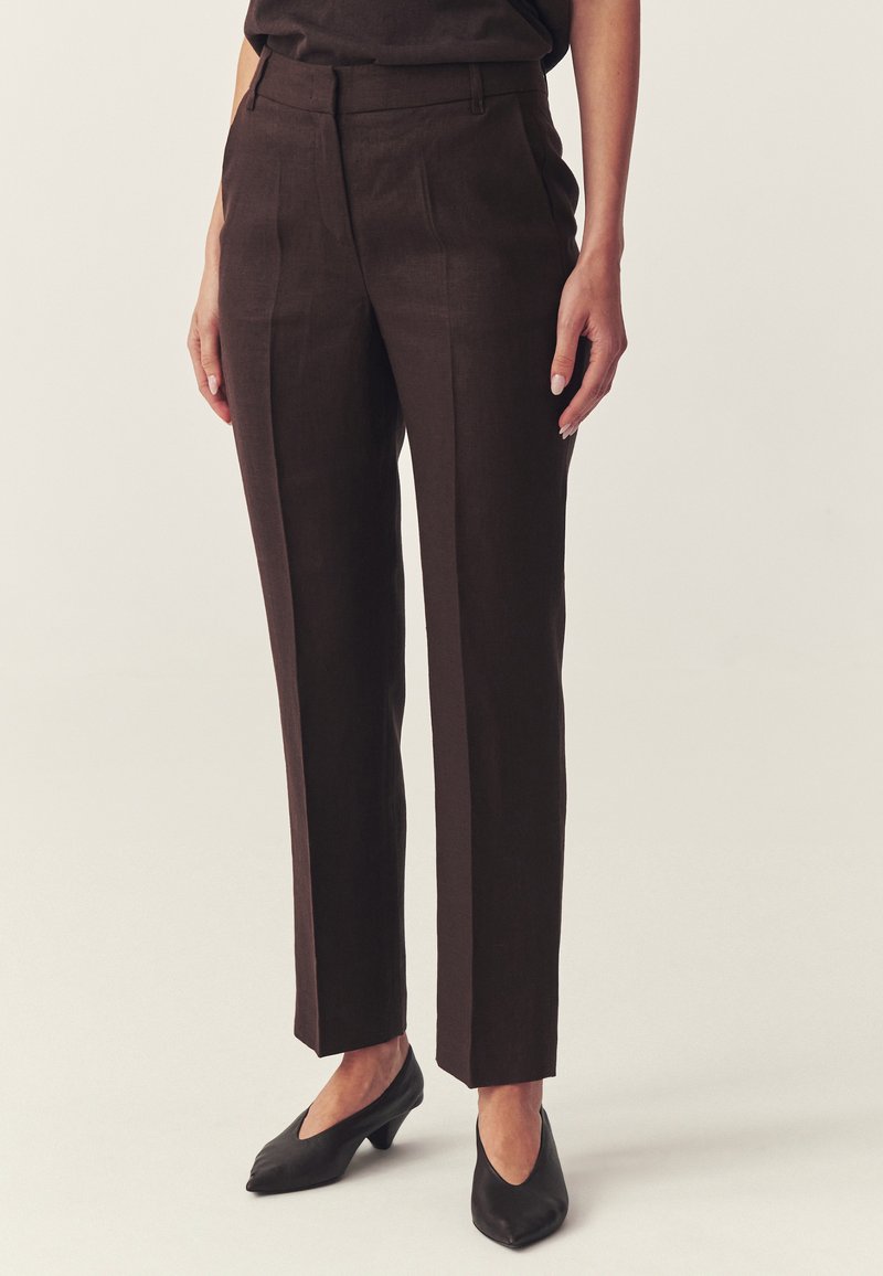 Femme portant un pantalon marron foncé taille haute à jambes droites avec des plis, associé à des chaussures noires à talons bas et bout pointu sur fond clair.