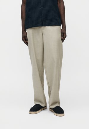 Άνδρας που φοράει φαρδιά μπεζ παντελόνια, μαύρα εσπαντρίγιες χωρίς κορδόνια και σκούρο μπλε υφασμάτινο πουκάμισο με κοντά μανίκια, στέκεται μπροστά σε απλό φόντο.