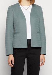 Světlomodrý blazer s texturovaným vzorem herringbone, otevřený vpředu, se dvěma bočními kapsami a dlouhými rukávy. Nositý přes bílý top.