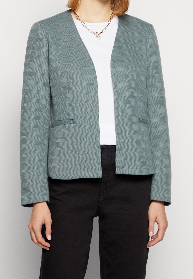 Světlomodrý blazer s texturovaným vzorem herringbone, otevřený vpředu, se dvěma bočními kapsami a dlouhými rukávy. Nositý přes bílý top.