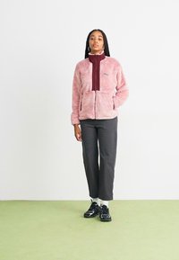 Lunefarvet fleecejakke med høj krave, lynlås foran og to sidelommer; parret med mørke bukser og sorte sneakers.