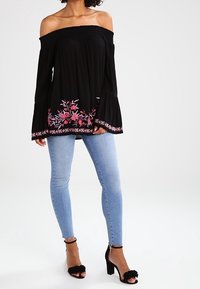 Top nero off-the-shoulder con ricami floreali in rosa e bianco, abbinato a jeans skinny azzurri chiari e sandali neri con tacco adornati.