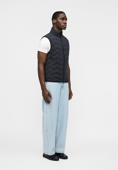 Άνδρας που φοράει μαύρο κοντό γιλέκο με ραβδώσεις πάνω από άσπρο πουκάμισο με κοντά μανίκια, ανοιχτό μπλε, φαρδιά τζιν παντελόνια και μαύρα παπούτσια, στέκεται μπροστά σε απλό λευκό φόντο.