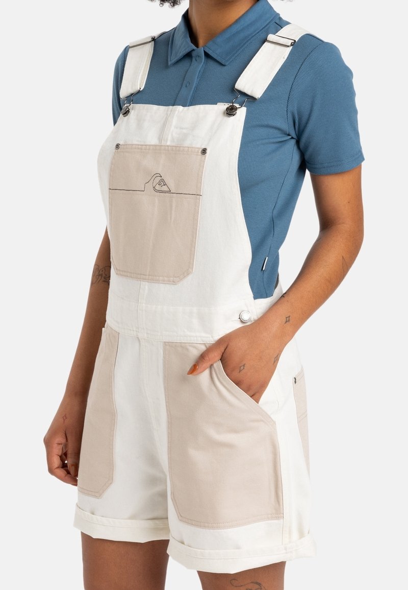 Salopette en coton blanc avec des patchs beige, bretelles réglables, poche frontale et poches latérales ; coupe décontractée avec ourlets retroussés.