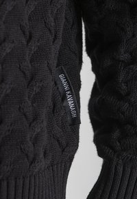 Suéter negro de punto con un patrón de trenzas, con un dobladillo de canalé. Se puede ver una etiqueta con "GIANNI KAVANAGH" en el lateral.