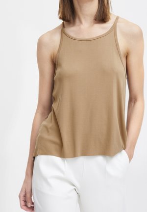 Femme portant un débardeur marron clair associé à un pantalon blanc, debout avec une main dans la poche devant un fond uni.