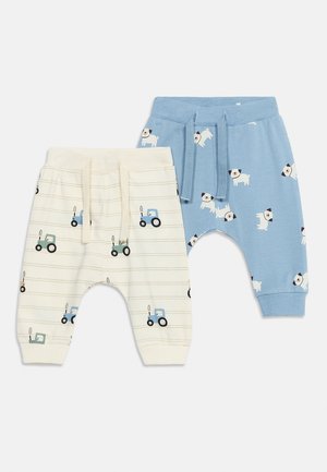 Deux pantalons pour bébé : un crème avec des imprimés de tracteurs bleus et verts, un bleu avec des imprimés de chiens blancs, tous deux avec ceinture à cordon de serrage.