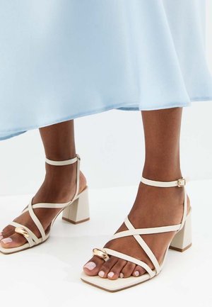 Donkere voeten met beige hoge hakken sandalen met enkelbandjes en gouden teenterringen, gecombineerd met een lichtblauwe midirok.