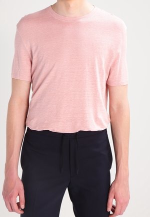 Mann trägt ein hellrosa kurzärmeliges T-Shirt, das in schwarze Kordelhosen gesteckt ist, und steht vor einem schlichten Hintergrund.