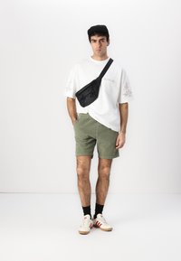 Schwarze Bauchtasche, getragen über einem weißen T-Shirt mit Blumenmuster, olivgrüne Shorts, schwarze Socken und beige Sneakers mit roten Streifen.