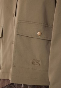 Κοντινό πλάνο τσέπης χακί μπουφάν με καπάκι ασφαλισμένο με μεταλλικό κουμπί και διακριτικό ραμμένο γεωμετρικό λογότυπο κάτω από την τσέπη.