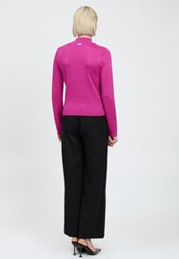 Persona con capelli biondi corti che indossa un top magenta aderente a maniche lunghe, pantaloni neri larghi e scarpe col tacco nere, in piedi di spalle.