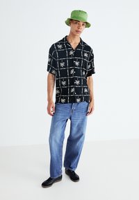 Camisa negra de manga corta con estampado de palmeras blancas, combinada con jeans holgados de color azul claro y zapatos negros. Un sombrero de cubo verde completa el look.