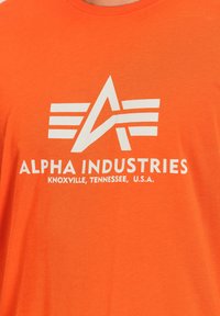 Camiseta de algodón naranja con un logotipo gráfico blanco que presenta letras estilizadas y tres franjas horizontales. El texto dice "ALPHA INDUSTRIES, KNOXVILLE, TENNESSEE, U.S.A."