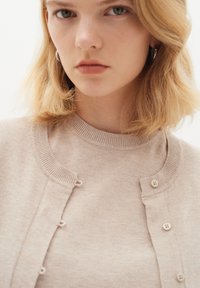Helles beige Cardigan mit rundem Halsausschnitt, kleinen Knöpfen und gerippten Kanten. Der Stoff wirkt weich mit einer feinen Strickstruktur.