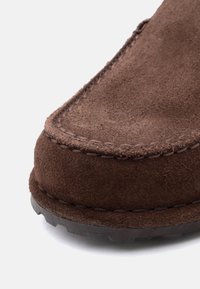 Καφέ μοκασίνι από σουέντ με εμφανείς ραφές και υφή στην άκρη της λαστιχένιας σόλας, σε λευκό φόντο.