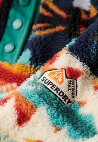 Fleece pullover with an abstract, colorful pattern in orange, blue, and cream, featuring a green button placket and a Superdry logo patch.