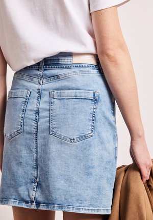 Jupe en denim bleu clair avec deux poches arrière portée avec un haut blanc, personne tenant une veste marron dans la main droite.