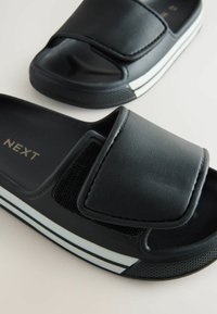 Sorte slidesandaler med en struktureret overdel, justerbar Velcro-strop og hvidstribede detaljer på sålen. Mærket 'NEXT' er synligt.