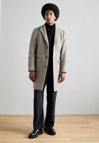 Abrigo de lana gris claro con solapas de muesca, cierre de botones y bolsillos laterales, combinado con un polo negro, pantalones de rayas y zapatos negros gruesos.