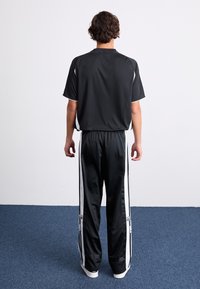 Una persona con i capelli corti e ricci è in piedi di fronte a una parete bianca, indossando un top nero a maniche corte e pantaloni sportivi neri e bianchi su un tappeto blu.