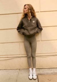 Sweatshirt en polaire vert olive foncé à zip quart et poche avant, leggings gris texturé, chaussettes blanches et baskets blanches avec des accents beiges.
