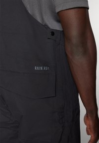 Svarte vanntette overaller med teksturert finish, med en sidelomme og merket "RAIN.RDY" i lys blått på forsiden.