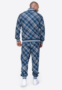 Mėlynas languotas treniruotinis kostiumas, pagamintas iš audinio su elastinėmis rankovėmis ir juosmeniu. Jame yra baltos ir tamsiai mėlynos raštai, sukurti patogumui ir judrumui.