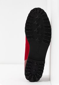 Semelle d'une botte rouge avec une bande de roulement noire, présentant des motifs en forme de croix et linéaires, positionnée verticalement sur un fond blanc.