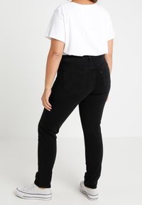 Jeans negros con un diseño ajustado y bolsillos traseros, combinados con una camiseta blanca. El modelo lleva zapatillas blancas.