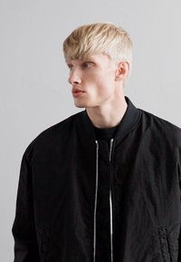 Jonge man met kort blond haar, gekleed in een zwarte zip-up jas, die naar links kijkt tegen een effen lichtgrijze achtergrond.