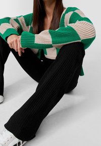 Gebreide oversized trui in groene en beige strepen, gecombineerd met zwarte geribde broek. Model zit en toont de pasvorm en textuur van de outfit.