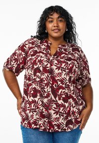Blusa de manga corta en crema claro con un patrón floral en rojo y marrón. Presenta un escote en V, tejido texturizado y un ajuste relajado.