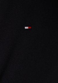 Schwarzer strukturierter Stoff mit einem kleinen rechteckigen Logo-Patch in Weiß, Schwarz und Rot, zentral platziert.