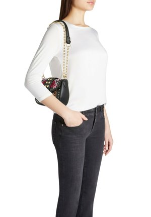 Borsa nera in pelle con tracolla a catena dorata, decorata con motivo floreale e dettagli borchiati, tenuta da una persona che indossa una camicia bianca e jeans neri.