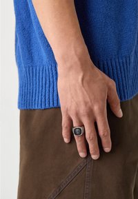 Sinine tekstuuriga sviiter ribatud alumise äärega, kantud pruunist kangast. Paremal käel on tume rõngas detailse embleemiga.