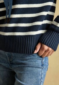 Tumesinise ja valge triibuline sveter ribadega mansettidega, valmistatud kudumaterjalist. Käsi toetub teksadele, näidates sobivat disaini.