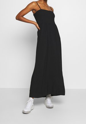 Zwarte maxi-jurk met smalle bandjes en elastisch lijfje, gedragen met witte hoge sneakers, model staat met één hand op de heup.