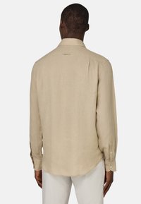 Camicia a maniche lunghe beige con una texture semplice, polsini abbottonati e un orlo leggermente curvato. Presenta un colletto standard e un dettaglio sulla spalla posteriore.