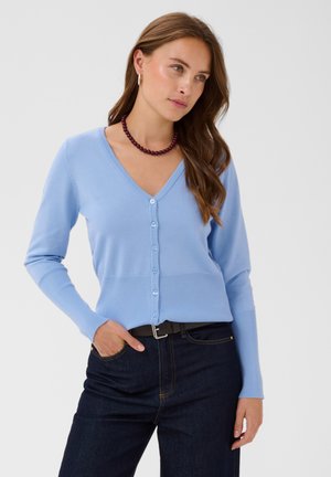 Femme aux longs cheveux bruns porte un cardigan bleu clair à col en V, un jean foncé, un collier de perles foncé et des créoles, la main dans la poche.