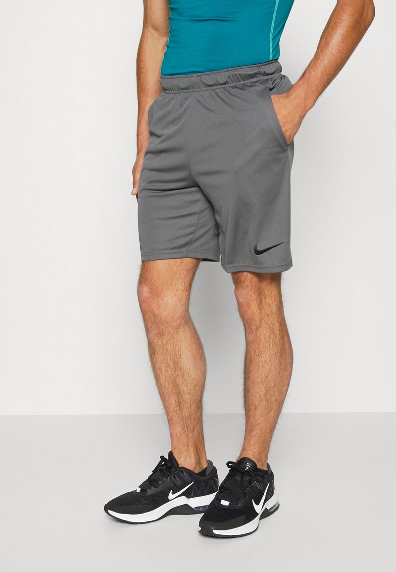 Šedé elastické kraťasy se bočními kapsami a černým logem Nike. Nosené s černými sportovními botami s bílými akcenty na neutrálním pozadí.