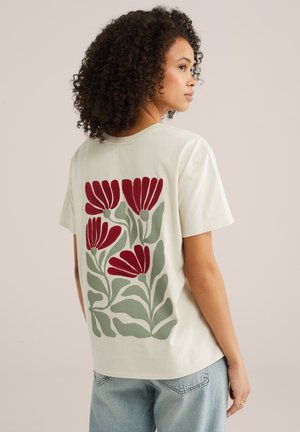 Femme aux cheveux bouclés portant un t-shirt blanc avec de grandes fleurs rouges et des feuilles vertes imprimées dans le dos.