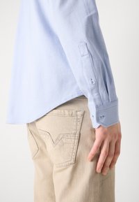 Ljusblå, texturerad långärmad skjorta med knapppdetaljer, bärs med beige byxor som har en karakteristisk fickform.