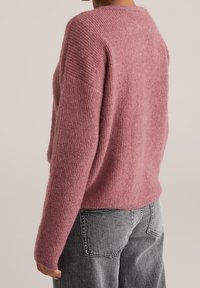 Světle růžový pletený svetr s volnějším střihem, kulatým výstřihem a žebrovanou strukturou. Kombinován se šedými džíny s vysokým pasem.