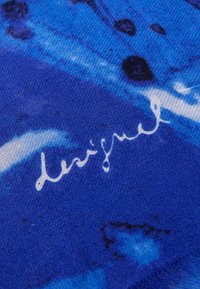 Tessuto blu con una gradazione di tonalità più scure e più chiare, con motivi astratti e un logo bianco scritto a mano nella sezione inferiore.