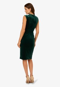 Zielona sukienka z aksamitnego materiału o dopasowanym kroju, bez rękawów, z detalami przy dekolcie z tyłu. W zestawieniu z cielistymi szpilkami.