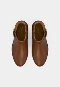 Smeđe umjetne gležnjače s glatkom teksturom, zaobljenim vrhom i detaljem s kopčom sa strane. Na etiketi piše "ANNA FIELD."