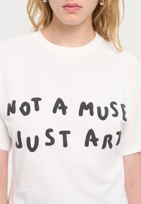 Närbild på en person som bär en vit skjorta med svart text som säger: "INTE EN MUSE, BARA KONST," och små hoopsörhängen.