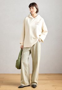 Κρέμα φούτερ με κουκούλα με γραφικά σχέδια, σε συνδυασμό με ανοιχτοπράσινα παντελόνια με φαρδιά ποδιά. Το μοντέλο κρατά μια πράσινη τσάντα με καπιτονέ σχέδιο και φορά μαύρα μυτερά παπούτσια.